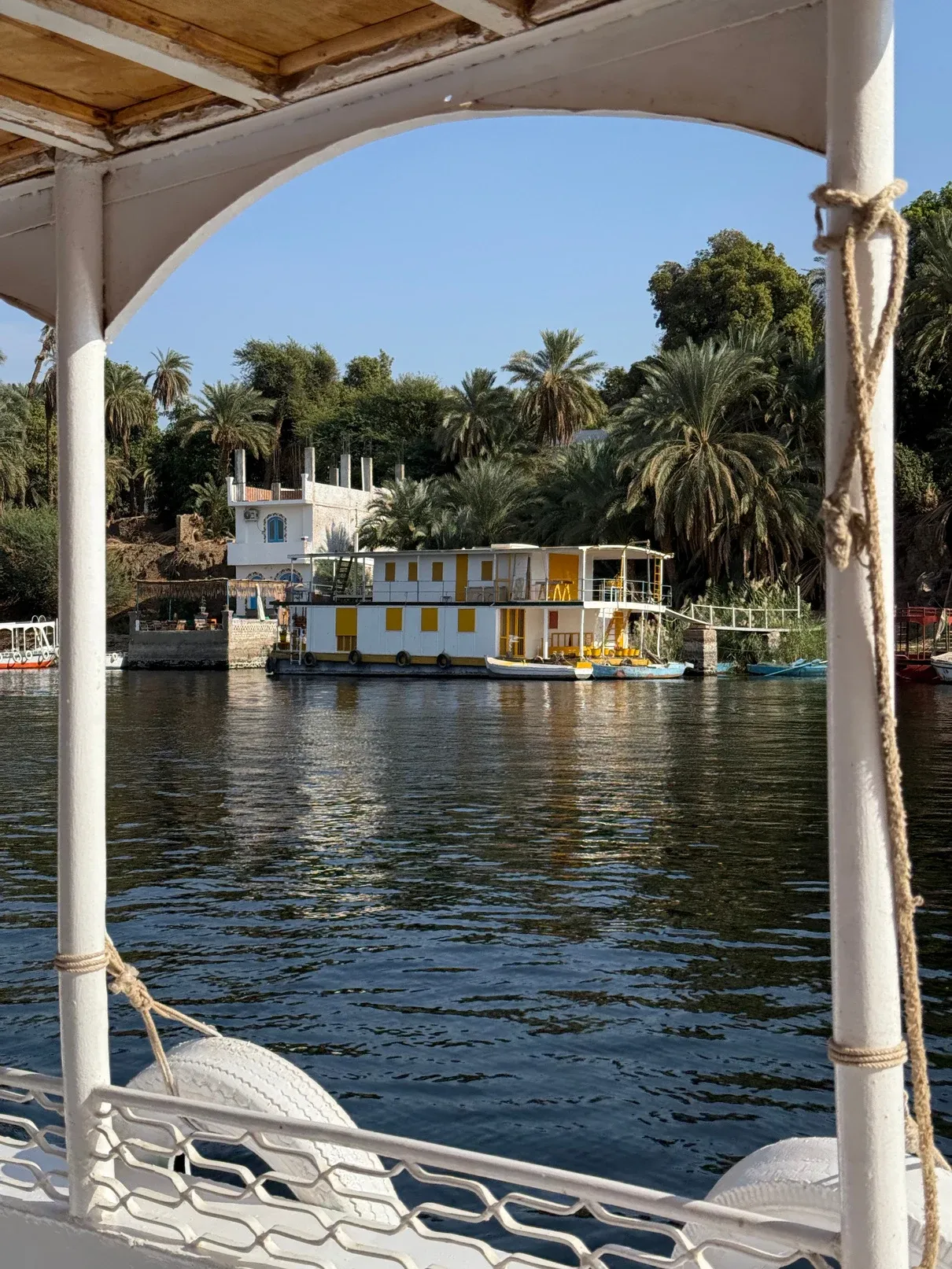 Blick durch den Holzbogen eines Bootsdachs auf ein gelbes Hausboot und Palmen am Nilufer.