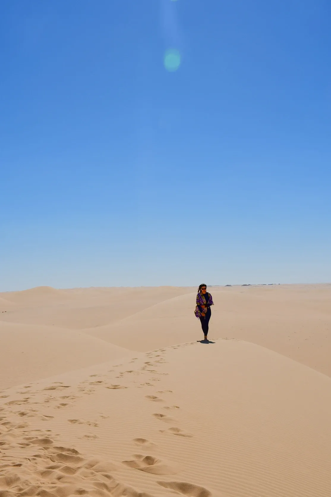Eine Frau steht allein auf dem Kamm einer Sanddüne, ihre Fußspuren ziehen sich durch den goldenen Sand.