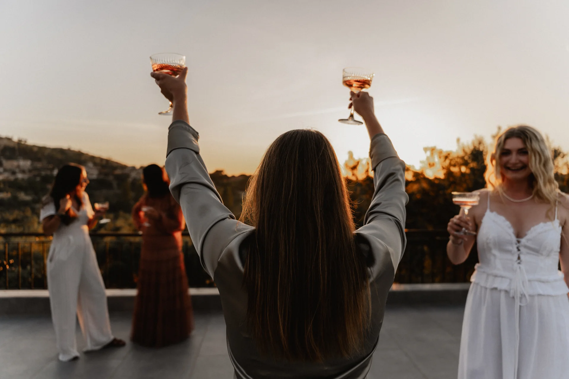Eine Frau reckt bei Sonnenuntergang auf einer Terrasse zwei Gläser in den Himmel, um sie herum stoßen Freundinnen an