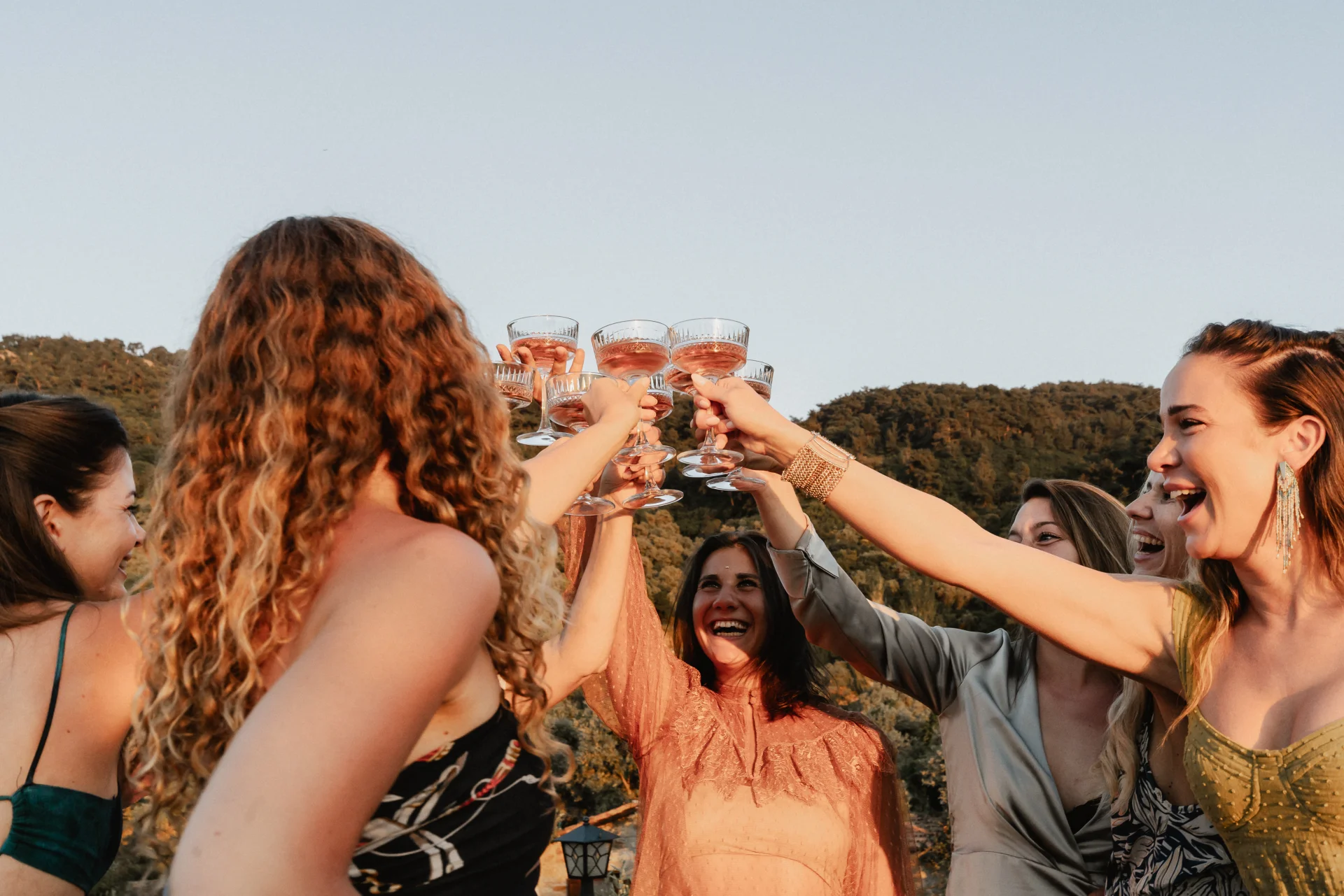 Frauen der Worldculture Travels Community stoßen bei Sonnenuntergang mit erhobenen Gläsern vor grünen Hügeln an