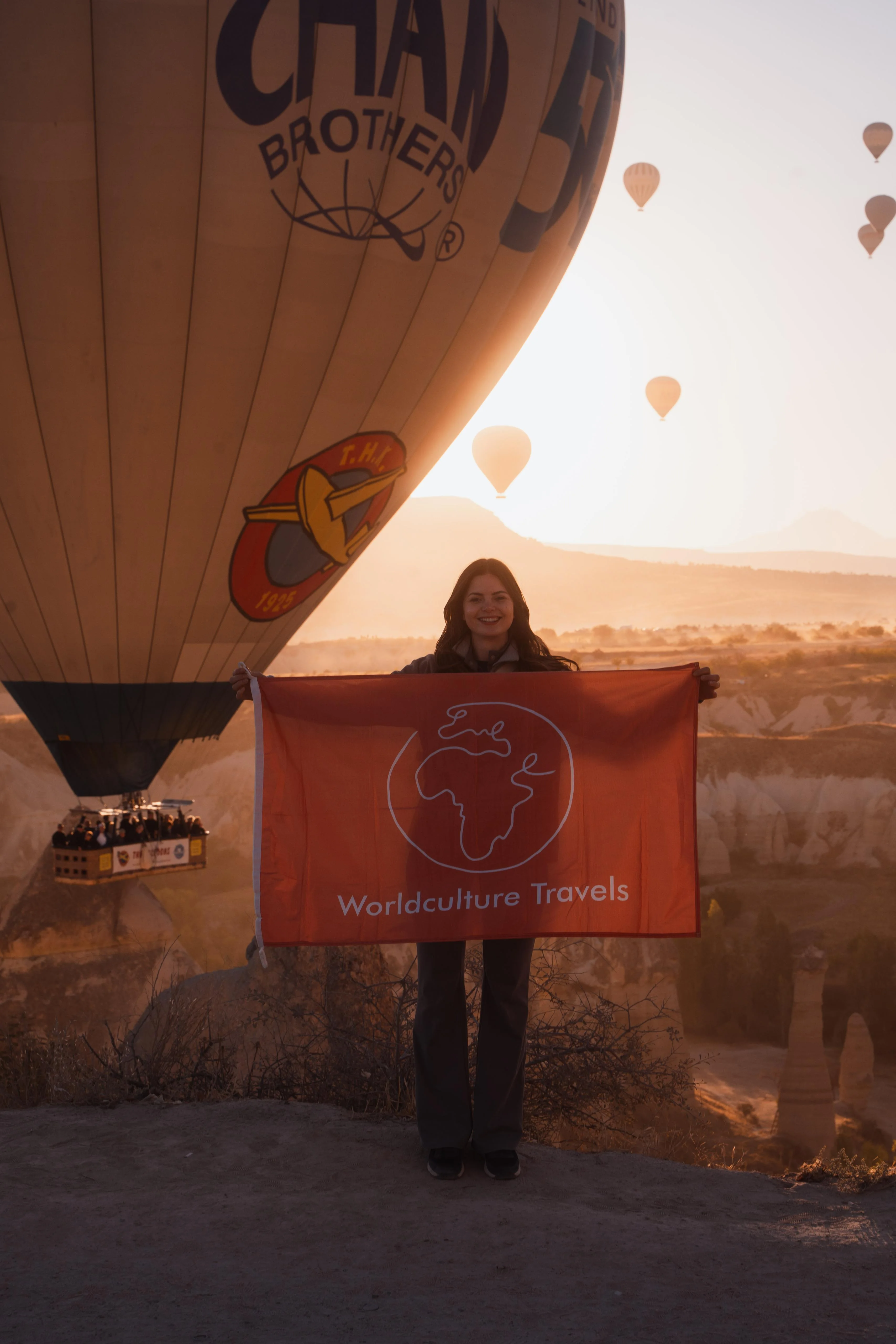 Selin hält bei Sonnenaufgang die orangefarbene Worldculture-Travels-Flagge vor Heißluftballons in Kappadokien